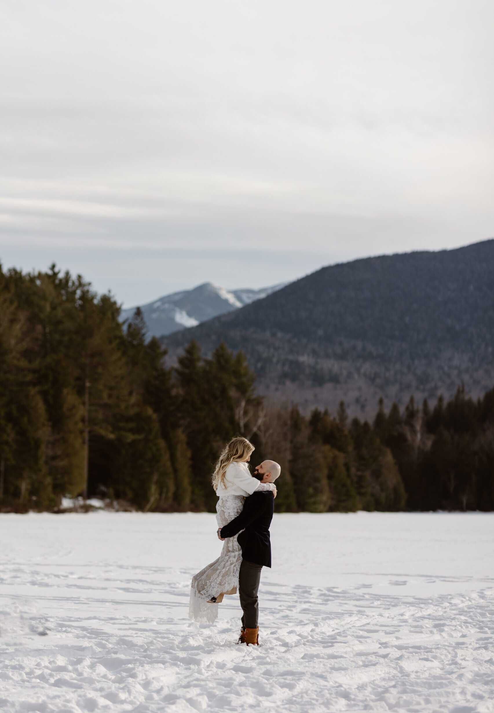 groom picks up bride on a frozen lake with snowy mountains behind them in the adirondacks