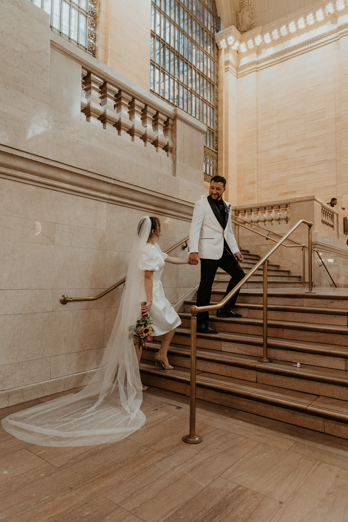 bride wearing short wedding dress with long veil and groom wearing white suit jacket with black pants walk together up the stairs in grand central station in new york city