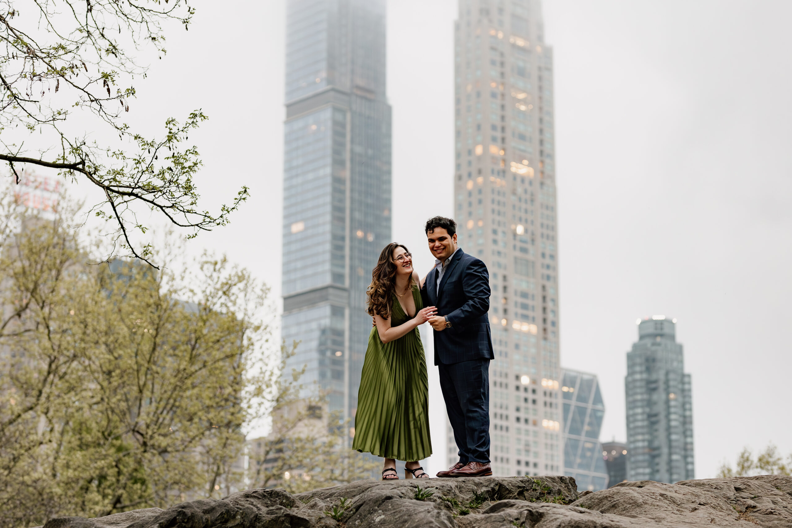 engaged couple smiling and laughing on top of boulders in central park, nyc with skyscrapers in the background