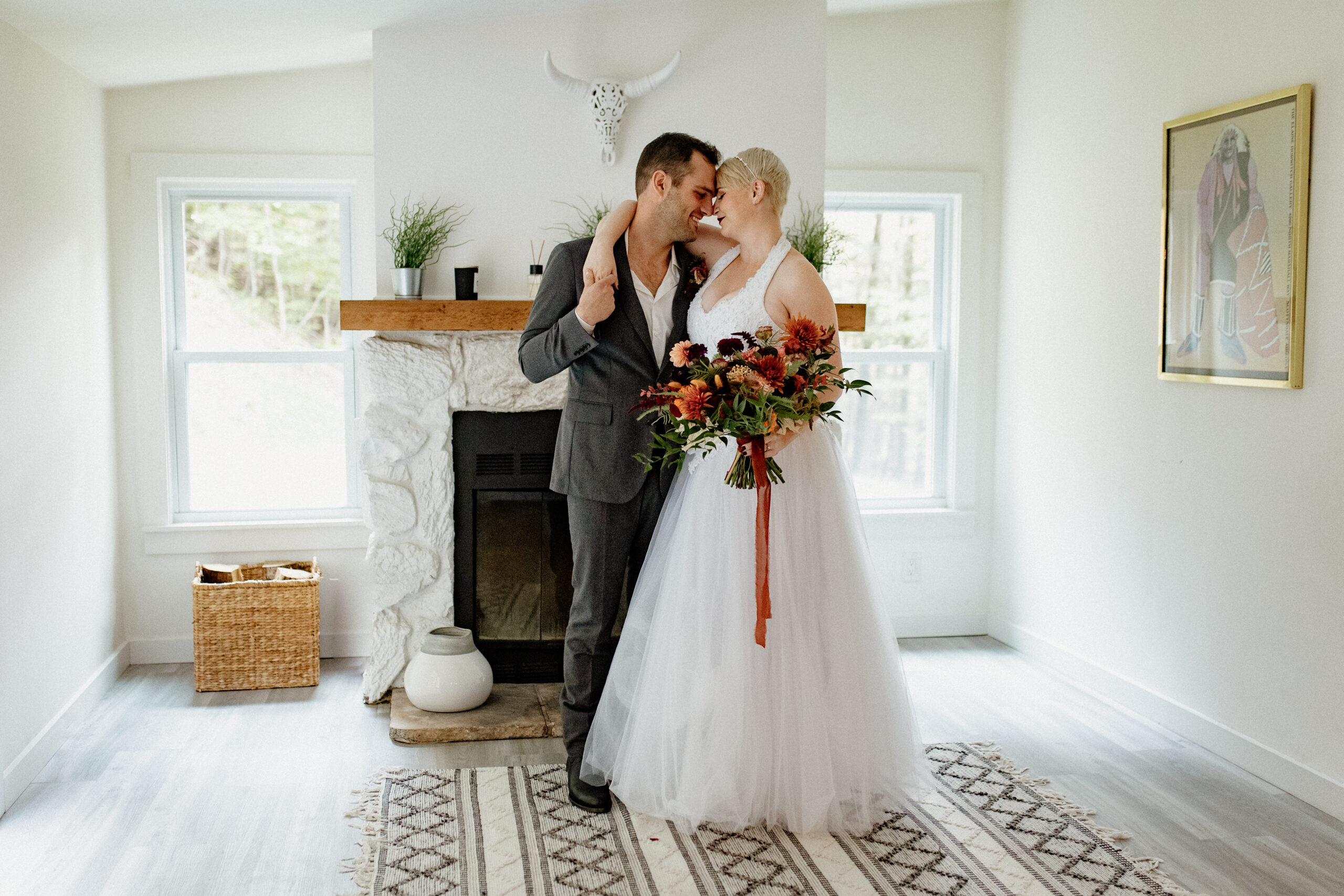 bride and groom hold onto each other in bright white room with boho decor, bride holds colorful bouquet