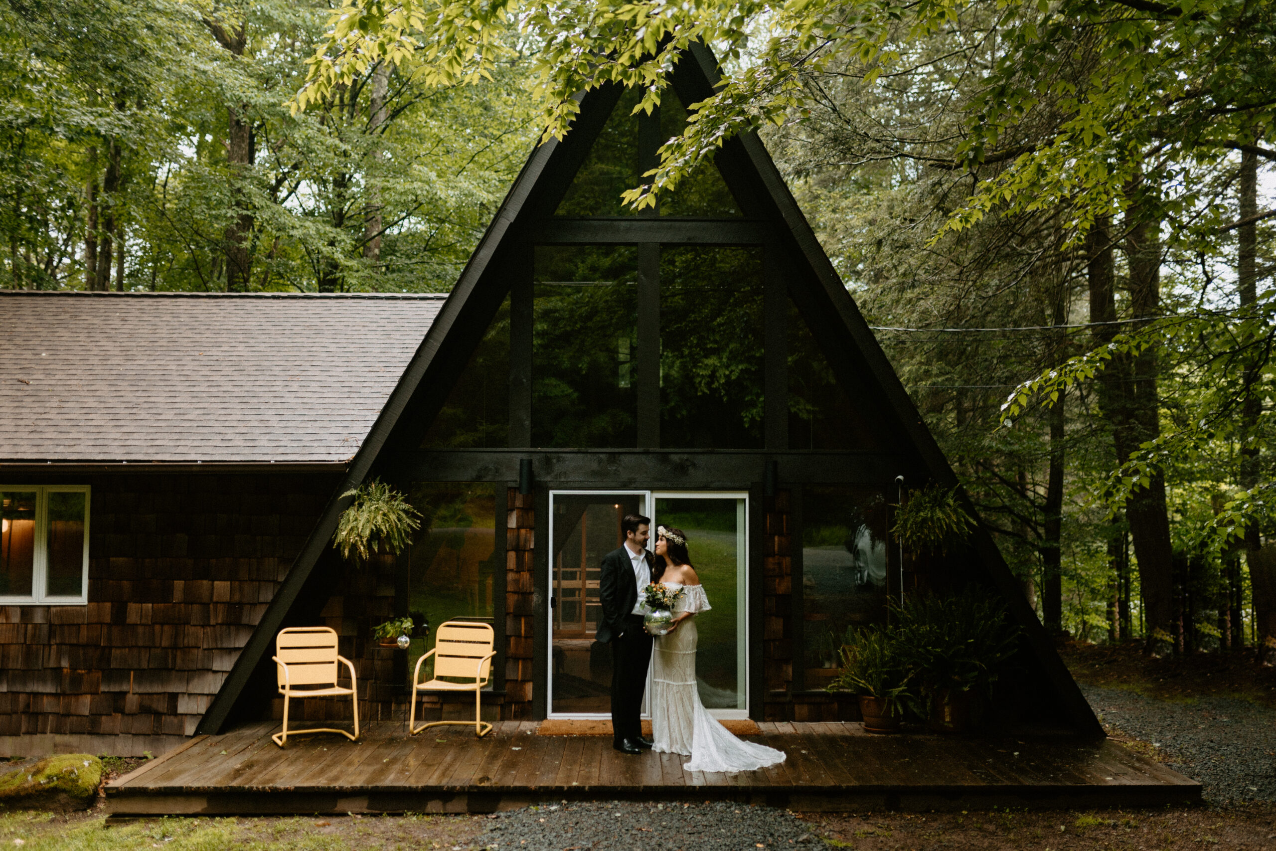 Eloping couple looks toward each other standing in front of an A-Frame cabin in the Poconos in the summer.